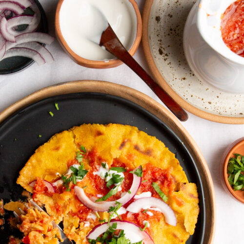 A black plate holds an Estrujadas Veracruzanas, a traditional Mexican dish from Tuxpan, topped with sliced red onions, cilantro, and red salsa. A fork rests alongside it. Nearby, bowls of creamy white sauce and chopped herbs accompany a mug with extra red salsa on a speckled plate.