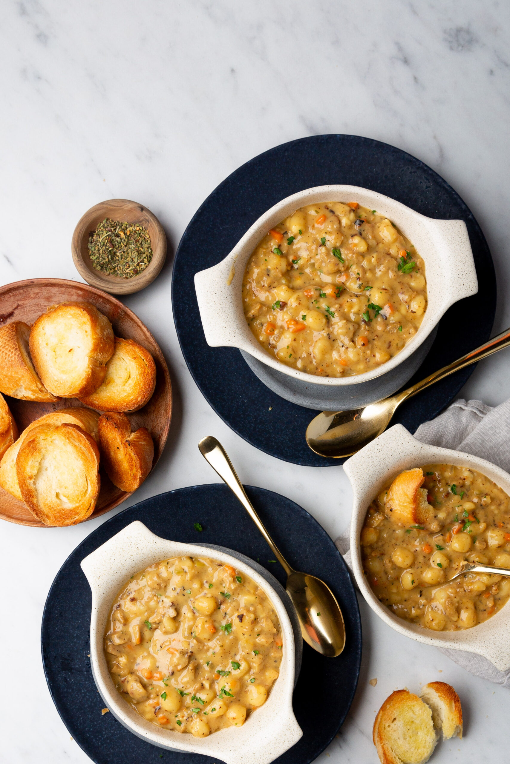 Three bowls of plant-forward, creamy soup garnished with herbs sit on dark plates. A gold spoon rests by each bowl. To the left, a wooden plate holds slices of toasted bread and a small bowl of dried herbs on a white marble surface.