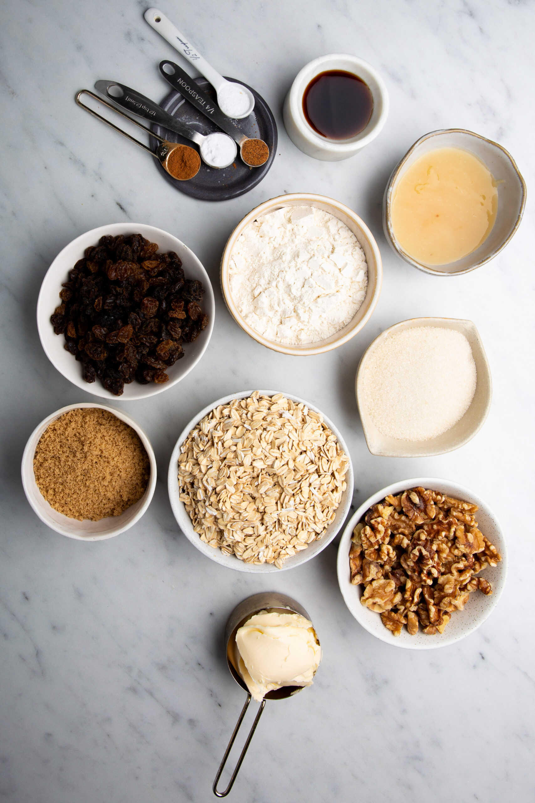 Various baking ingredients are arranged on a marble surface: small bowls hold raisins, flour, sugar, oats, brown sugar, walnuts, and a creamy mixture for vegan oatmeal cookies. Measuring spoons contain spices and baking soda. A small dish holds vanilla extract, with butter portioned in a measuring cup.