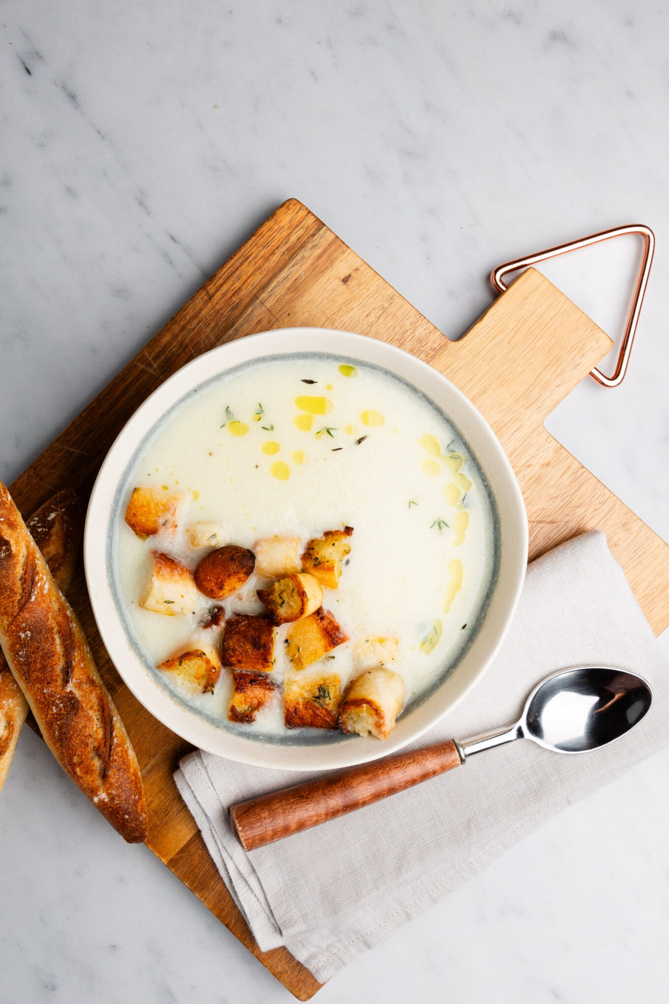 A bowl of vegan cauliflower soup garnished with croutons, drizzled with olive oil, and sprinkled with herbs sits on a wooden board. A crusty baguette is on the side, along with a spoon resting on a folded napkin. The setting is on a light marble surface.