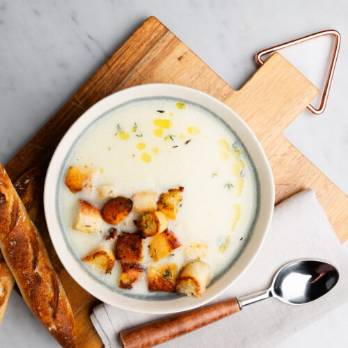 A bowl of vegan cauliflower soup garnished with croutons, drizzled with olive oil, and sprinkled with herbs sits on a wooden board. A crusty baguette is on the side, along with a spoon resting on a folded napkin. The setting is on a light marble surface.
