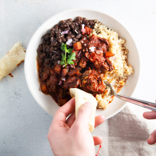 A white bowl filled with rice, black beans, and chile colorado stew topped with cilantro sits on a light surface. A person’s hands hold a fork and a piece of tortilla, about to scoop up food. A beige napkin with an orange edge is nearby.