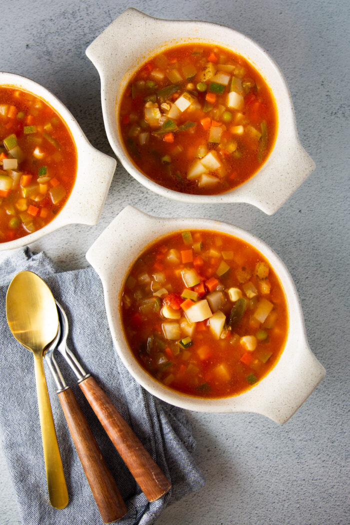 Three bowls of Sopa De Verduras soup with spoons on a napkin.