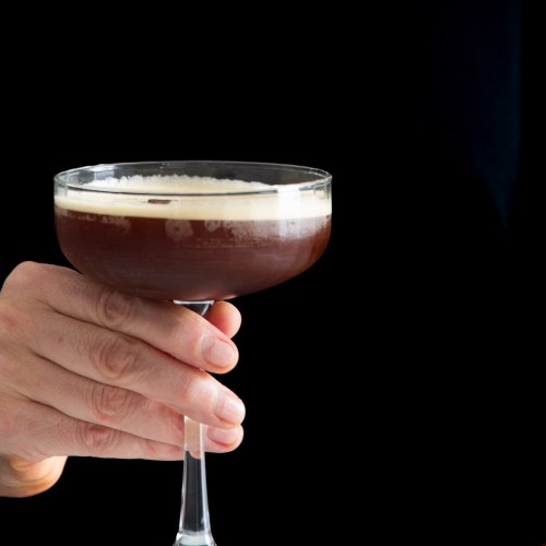 A hand holds a coupe glass filled with a dark espresso martini, topped with frothy crema. The black background highlights the drink and hand, evoking the bold allure of a shaken carajillo, perfect for fans of Mexican coffee cocktails.