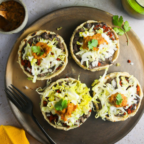 A round wooden plate holds four homemade sopes topped with refried beans, shredded lettuce, sour cream, salsa, and cilantro. A fork, knife, yellow napkin, small bowl of salsa, green drink, and jug sit nearby on a light surface.