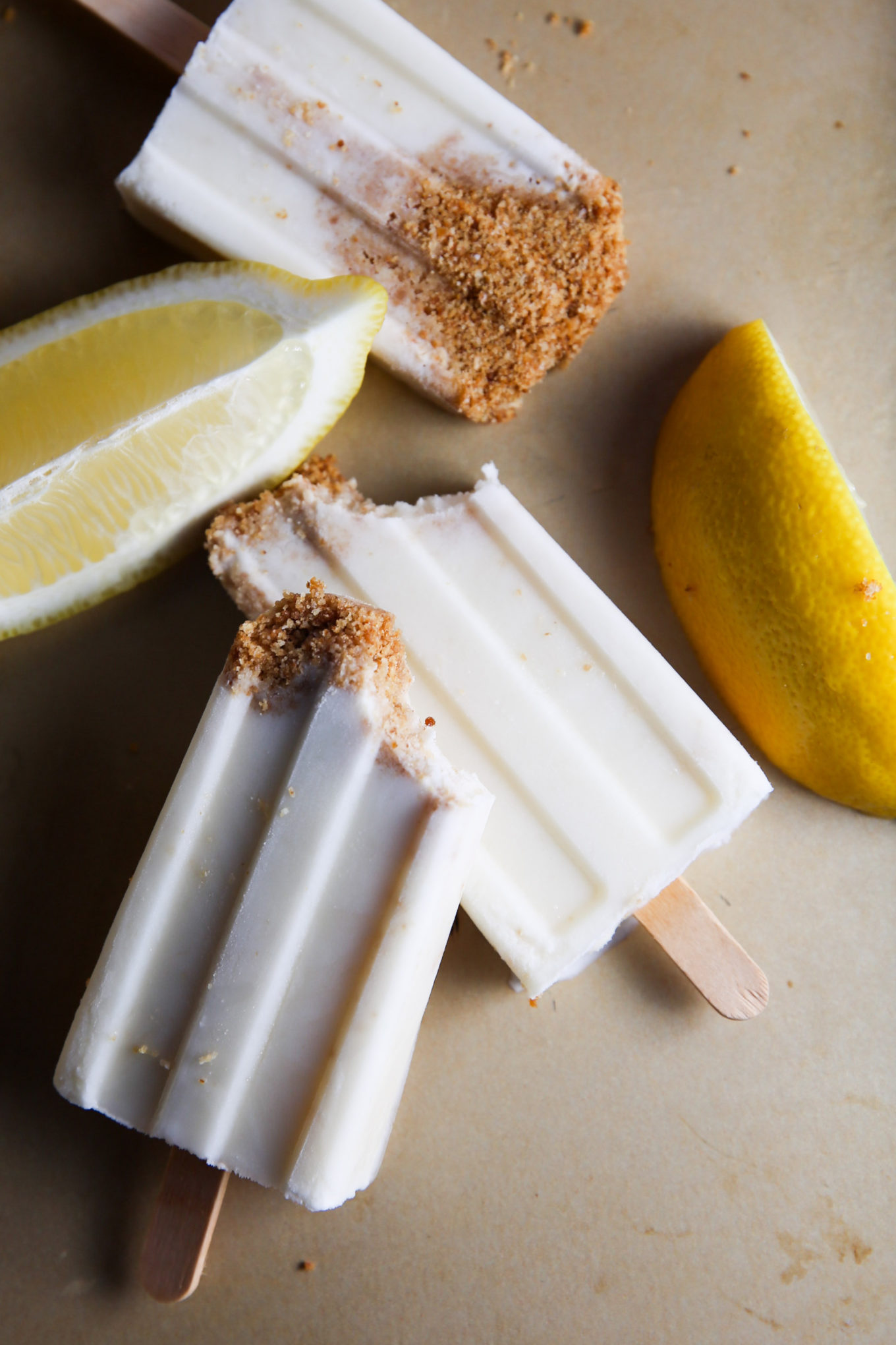 Three lemon pie-flavored paletas with brown sugar crust on a beige surface, accompanied by fresh lemon wedges. The paletas are partially visible and rest vertically.