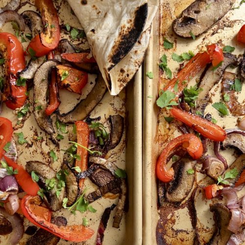 Two sheet pans filled with roasted vegetables, including red bell peppers, red onions, and portobello mushrooms. The seasoned veggies are garnished with fresh cilantro. A slightly charred flour tortilla is placed in the top left corner, partially covering some vegetables—perfect for a fajitas recipe.