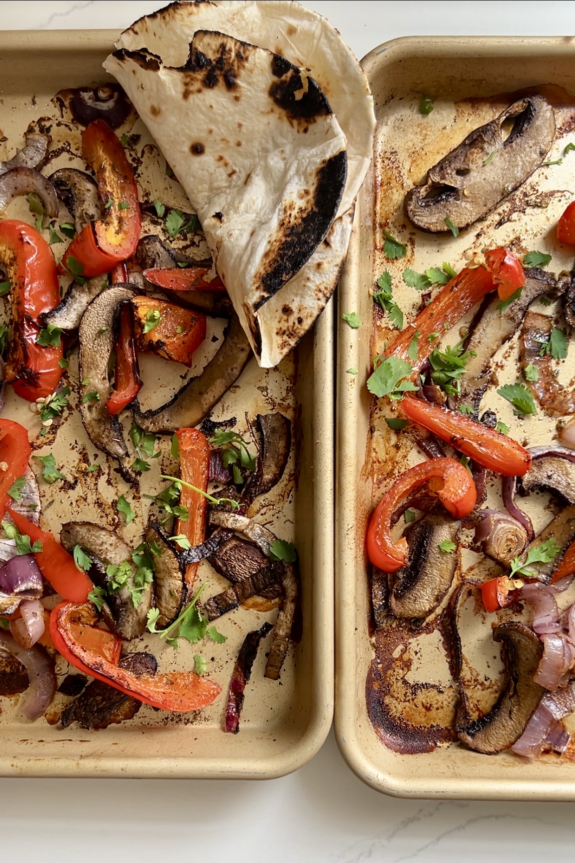 Two sheet pans filled with roasted vegetables, including red bell peppers, red onions, and portobello mushrooms. The seasoned veggies are garnished with fresh cilantro. A slightly charred flour tortilla is placed in the top left corner, partially covering some vegetables—perfect for a fajitas recipe.