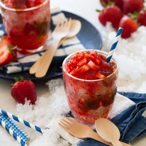 Two glasses of strawberry raspado surrounded by crushed ice, whole strawberries, and wooden utensils on a white surface, with blue-striped straws adding a touch of color.