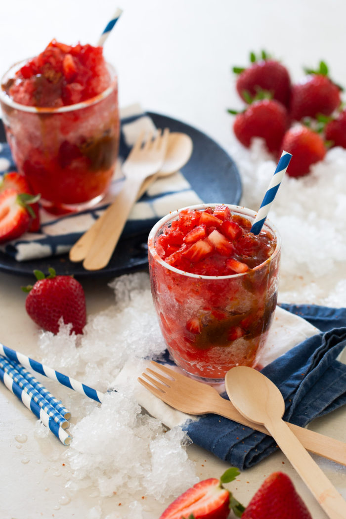 Two glasses of strawberry raspado surrounded by crushed ice, whole strawberries, and wooden utensils on a white surface, with blue-striped straws adding a touch of color.