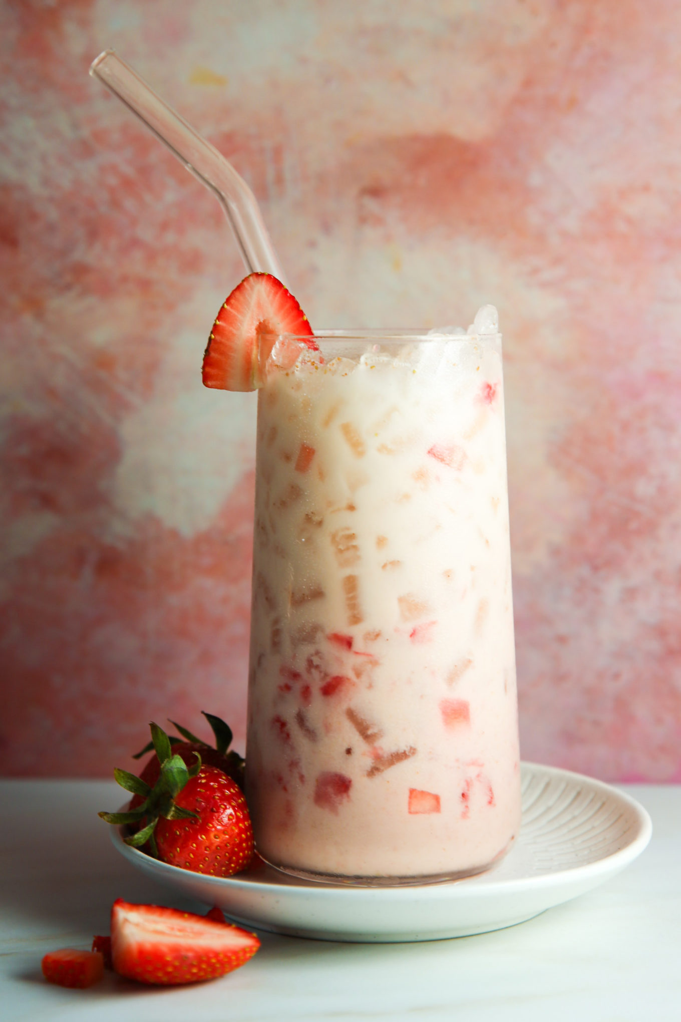 A refreshing strawberry horchata in a clear glass, filled with ice and diced strawberries, topped with a whole strawberry on the rim. The drink is served with a glass straw, on a light plate