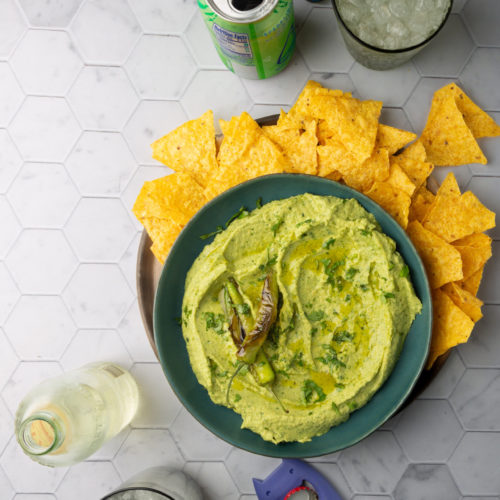 A bowl of avocado hummus topped with grilled jalapenos, surrounded by corn tortilla chips on a hexagon-patterned surface. Nearby are drinks, including a green can and a clear bottle, both served in glasses with ice. A juiced lime half and a blue shark-shaped bottle opener complete the scene.
