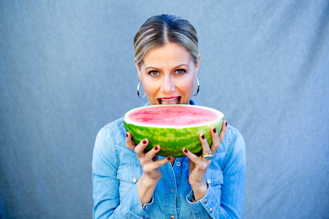 A woman with blonde hair in a ponytail, wearing a light denim shirt, holds a large slice of watermelon near her mouth, about to take a bite. She has hoop earrings and dark nail polish&mdash;perfect inspiration for your next chia recipe or bowl of overnight oats.