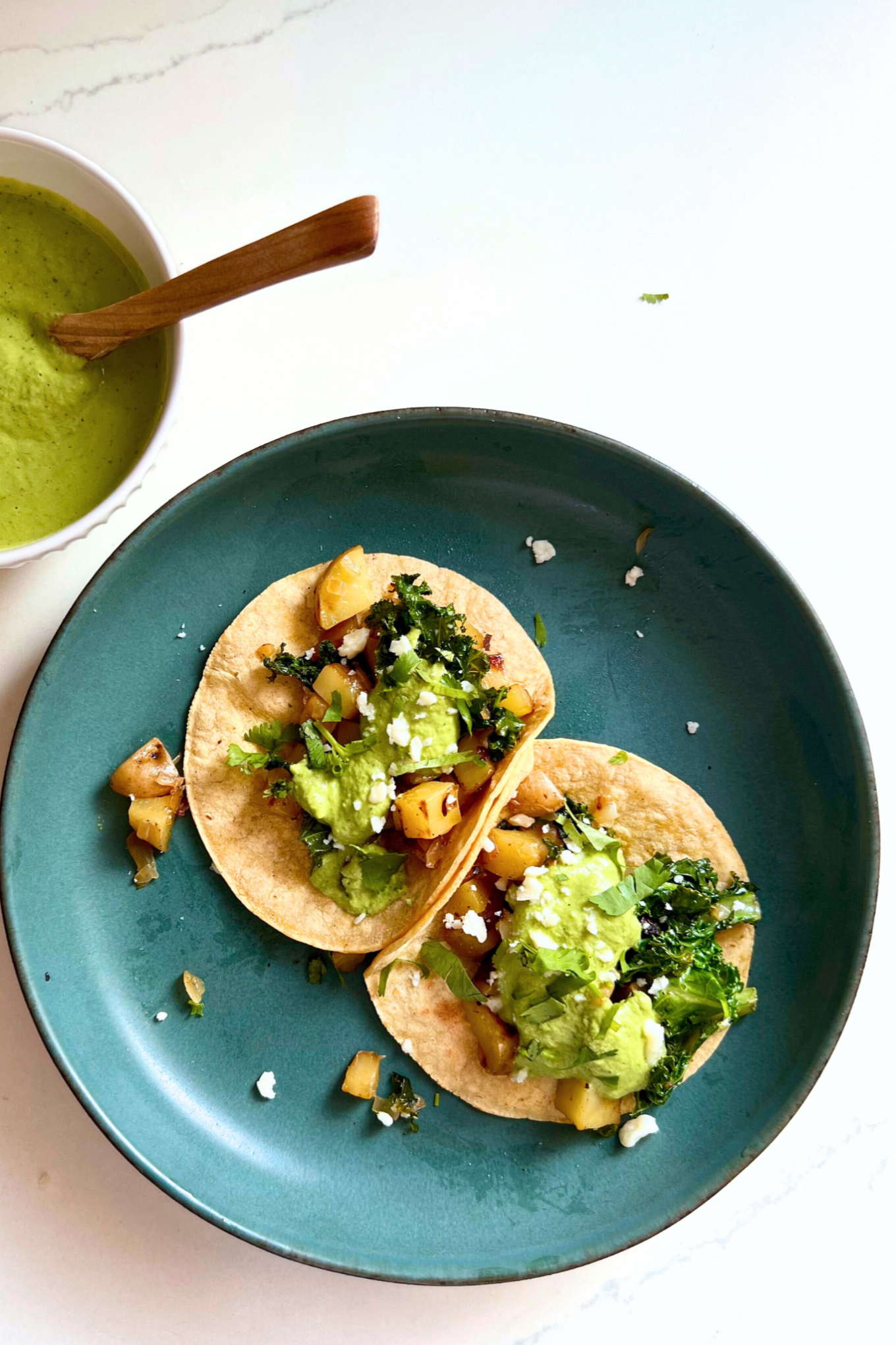 Two soft tacos filled with saut&eacute;ed vegetables, kale, and crumbled cheese sit on a blue-green plate. Nearby, a bowl of cilantro Bitchin Sauce with a wooden spoon is placed on a white surface. Small bits of taco filling and cheese crumbs surround the tacos on the plate.