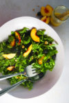A white bowl brimming with a vibrant arugula salad showcases peach slices, blackberries, and nuts, with tongs resting inside. Nearby, a jar of dressing with a spoon sits beside extra peach slices. The early morning sun casts soft shadows across the scene.