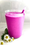 A vibrant purple Dragon Fruit Smoothie in a transparent glass with a clear straw is placed on a white marble surface. Three small white daisies with yellow centers and green leaves rest beside the glass, adding charm to this healthy and easy treat. The background features white subway tiles.