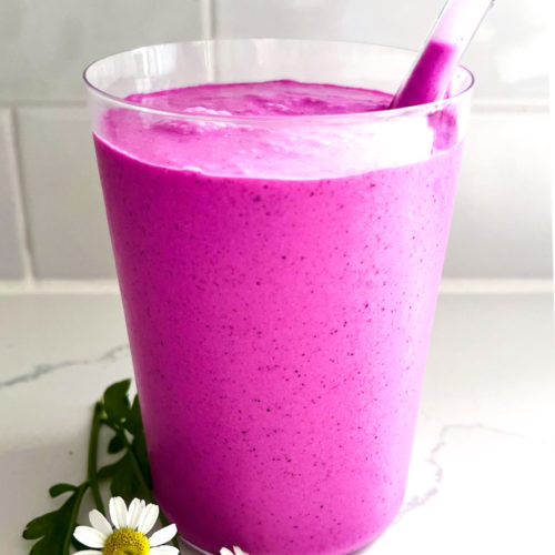 A vibrant purple Dragon Fruit Smoothie in a transparent glass with a clear straw is placed on a white marble surface. Three small white daisies with yellow centers and green leaves rest beside the glass, adding charm to this healthy and easy treat. The background features white subway tiles.