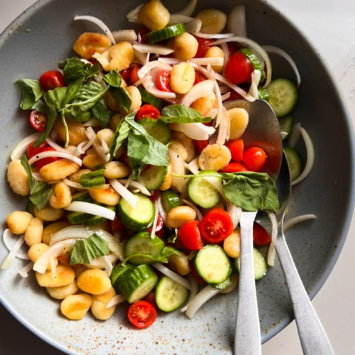 A vibrant bowl of pan-fried gnocchi salad with sliced cucumbers, cherry tomatoes, onions, and fresh basil leaves. Two silver spoons rest inside the bowl on a speckled, light surface. This colorful and fresh panzanella-inspired dish showcases a delightful mix of textures.