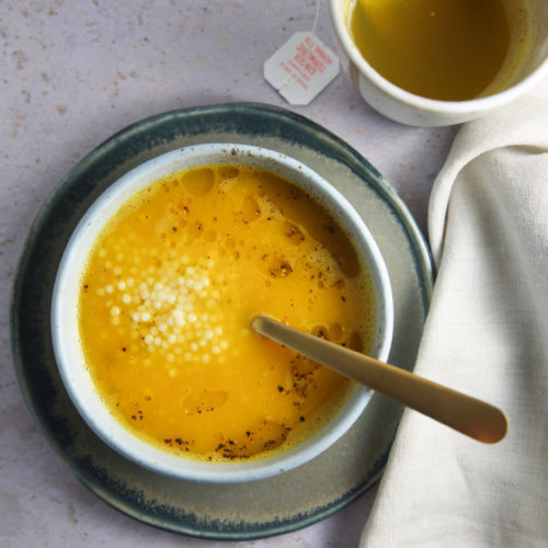 A bowl of yellow pastina soup with a sprinkle of white tapioca pearls, served in a white bowl on a greenish-grey plate with a gold spoon. Next to it is a white cup containing a yellow liquid and a used tea bag tag. A cream-colored cloth napkin is partially visible on the right.