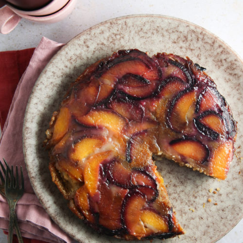A round plum upside-down cake with a slice removed sits on a speckled ceramic plate. The cake showcases a glossy, caramelized plum topping in shades of orange and purple. Nearby lie a red napkin, two forks, and stacked ceramic cups with a dusky pink hue on a light, textured surface.