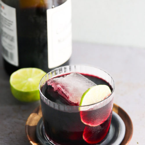 A glass filled with red wine, garnished with a lime slice and a large ice cube, sits atop a wooden and ceramic coaster. Behind the glass is a bottle of Tinto de Verano with an attached label, and a halved lime is placed next to the bottle on a light gray surface.