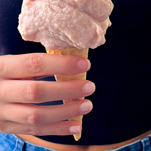 A person holds an ice cream cone with a large scoop of pink tofu ice cream. They are wearing a black top and blue jeans with a visible button. The person's nails are neatly trimmed and without polish, holding the cone with a gentle grip. The background is not visible.