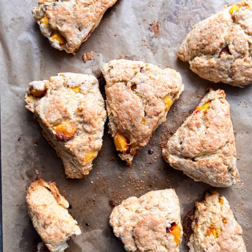 A baking sheet covered with parchment paper displays eight freshly baked, delicious peach scones, golden brown and studded with chunks of fruit. The scones have a rustic, homemade appearance, and some crumbs are scattered on the parchment surface. This easy recipe guarantees a delightful treat.
