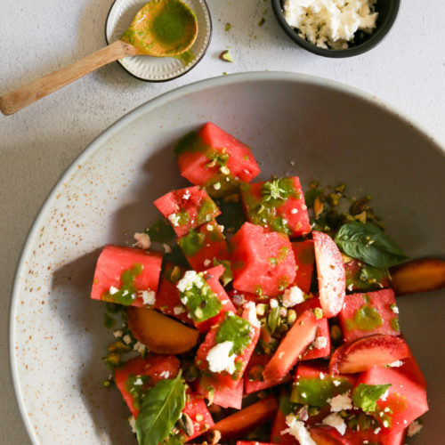 A bowl of watermelon salad with pieces of watermelon and peach, sprinkled with crumbled feta cheese, basil leaves, and a basil vinaigrette. Nearby, a small dish with green powder and a wooden spoon, a black bowl of crumbled feta, and a jar of basil vinaigrette are arranged on the gray surface.