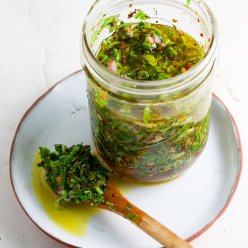 A glass jar filled with green chimichurri sauce, placed on a white plate with a red rim. A wooden spoon covered in the sauce is resting on the plate next to the jar, showcasing a mixture of chopped herbs and seasoning. This authentic chimichurri recipe shines against the textured, white background.