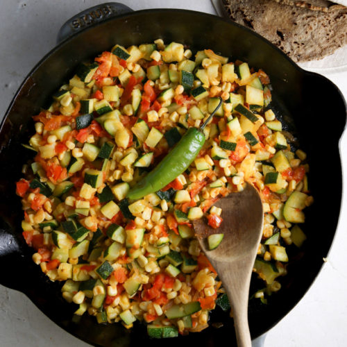 A black Staub pan sits on a white countertop, filled with a colorful mix of chopped vegetables including zucchini, tomatoes, and corn. A green chili pepper rests on top of the vegetables, and a wooden spoon lies in the pan. In the background, there is a small bowl of salt and a stack of tortillas.