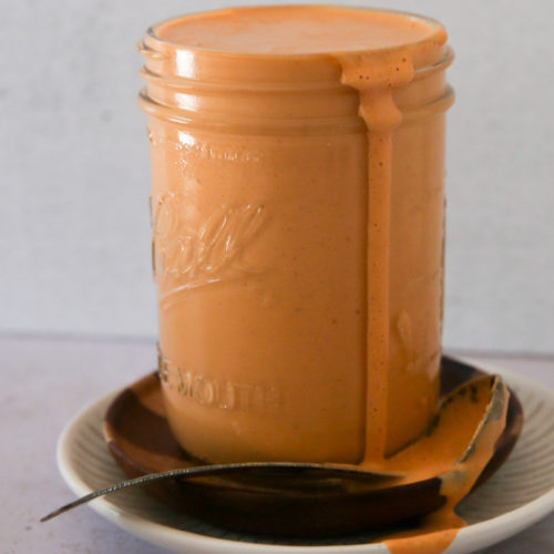 A jar filled with creamy, orange-colored chipotle crema overflows slightly, with the sauce dripping down the side. The jar is placed on two stacked saucers, one dark and one light. A spoon with sauce remnants lies across the saucers. The white background enhances the sauce's vibrant color.