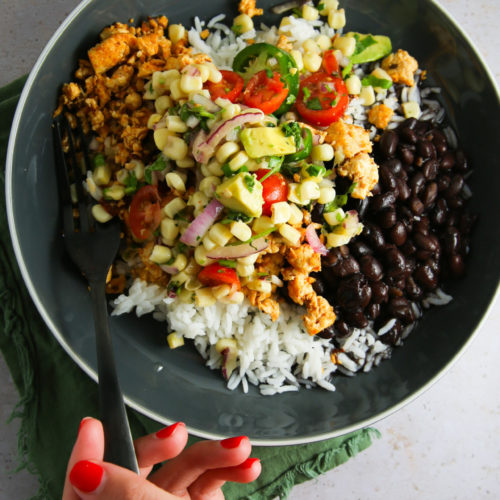 A black bowl filled with cooked ground tofu, black beans, white rice, and a tomato, red onion, avocado, and corn salsa is held by a hand with red nail polish. This vegan burrito bowl rests on a green napkin on a textured gray surface. The person's hand holds a fork dipped into the dish.