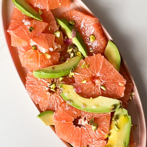 An oval copper platter holds an Avocado Grapefruit Salad with grapefruit slices and avocado wedges, sprinkled with crushed pistachios and drizzled with shallot vinaigrette. The bright colors are artfully arranged for a fresh, vibrant presentation.