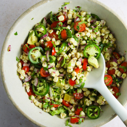 A bowl of corn salad with sliced jalapeños, cherry tomatoes, diced red onion, and chopped cilantro. Creamy avocado adds a delightful touch. A serving spoon rests in the bowl. The colorful mix contrasts against the vibrant greens and reds on a white surface.