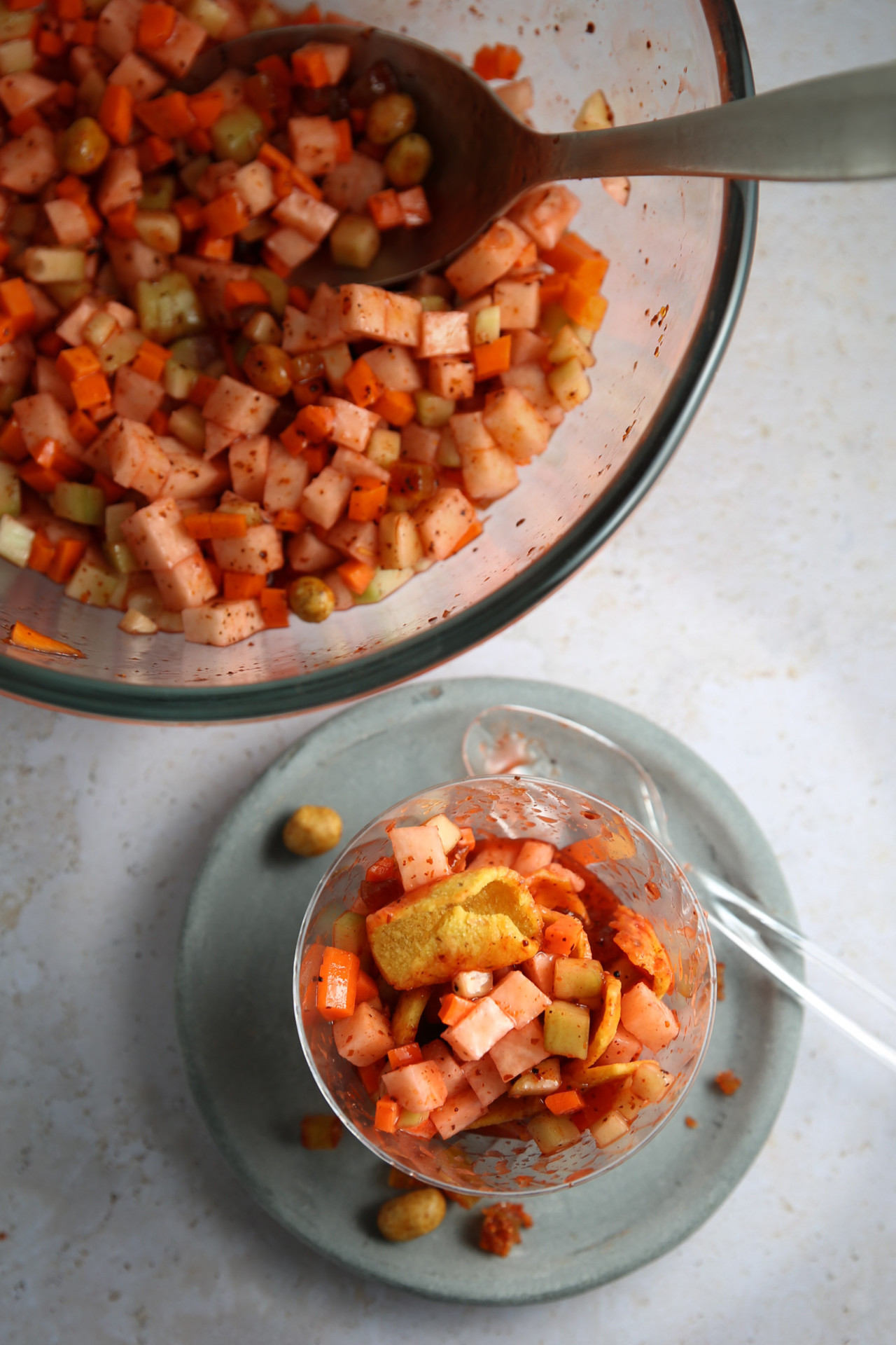 A close-up of a mixed vegetable salad in a clear glass bowl on a light grey plate with a transparent fork. The salad includes diced vegetables such as carrots, apples, and cucumbers, seasoned with spices—an ultimate party snack. A large bowl of the same salad and a serving spoon are visible in the background.