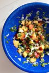 A vibrant Peach Pico De Gallo salsa in a blue speckled bowl on a white marble surface. This colorful 5-minute creation features diced peaches, chopped jalapeños, onions, and cilantro, blending hues of red, green, and white for a fresh and enticing appearance.