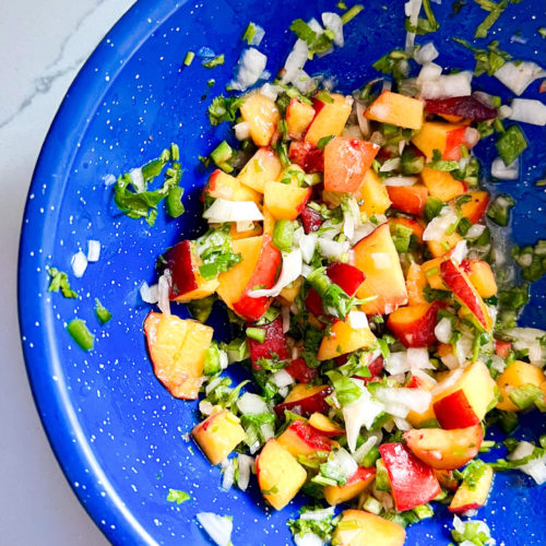 A vibrant Peach Pico De Gallo in a speckled blue bowl on a marble surface. The mix includes diced peaches, chopped cilantro, onions, and jalapeños, showcasing a colorful blend of orange, green, and white. Finely chopped ingredients create an appetizing dish that's perfect for any occasion.