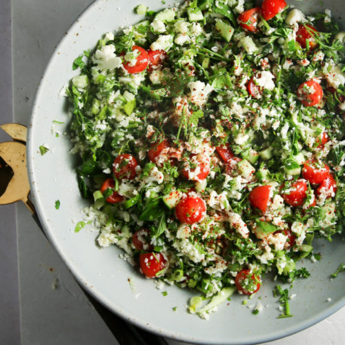 A large white bowl filled with a vibrant cauliflower tabbouleh sits on a light gray surface. This healthy recipe features cherry tomatoes, cucumber pieces, and chopped greens. A serving fork and spoon with black and bronze handles rest elegantly beside the bowl on the table.
