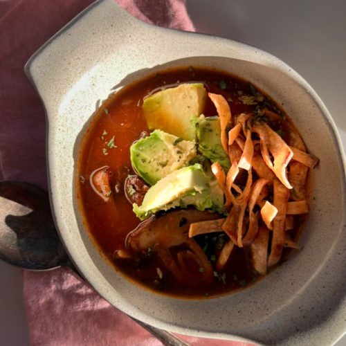 A bowl of Mexican Mushroom Soup sits on a pink cloth napkin. The soup features a rich, red chipotle-infused broth filled with chunks of avocado, crispy tortilla strips, and mushrooms. The bowl is made of white ceramic, and a silver spoon with a decorative handle rests to the side. The image is lit by natural light.