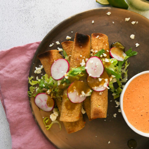 A wooden plate showcases three crispy taquitos topped with green salsa, sliced radishes, lettuce, and crumbled cheese. A pink napkin lies beneath the tacos. Beside them, a small bowl of chipotle dipping sauce and a glass with a lime wedge on the rim rest on a white surface.