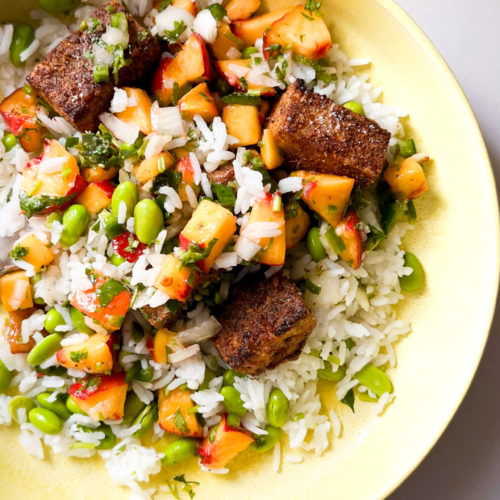 A vibrant rice bowl featuring fresh, colorful diced vegetables like edamame and bell peppers. Cubes of Mexican-spiced tofu are scattered on top, and the dish is garnished with chopped herbs. Finely mixed ingredients create an appetizing meal bursting with flavor.