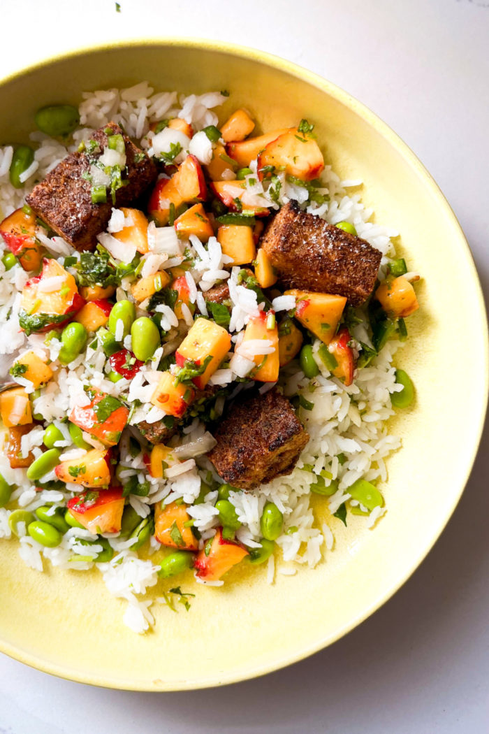A yellow bowl filled with white rice, green edamame beans, Peach Salsa, chopped onions, herbs, and browned cubes of Mexican-Spiced Tofu&mdash;all arranged on a white surface with natural lighting highlighting the fresh ingredients.