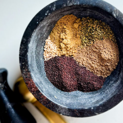 A top-down view of a black mortar containing five different ground spices arranged in sections, including Za'atar. The spices include beige, greenish, light brown, and dark reddish-brown powders. In the background, there is a pestle and two nested gold measuring spoons on a white surface, perfect for crafting an authentic recipe.