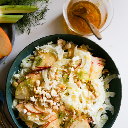 A fresh, vibrant apple salad sits in a green bowl, featuring thinly sliced fennel, apple slices, potato pieces, crumbled feta cheese, and hazelnuts. To the side is a glass jar containing a vinaigrette dressing and a spoon. Fresh fennel bulbs and sprigs are also partially visible.