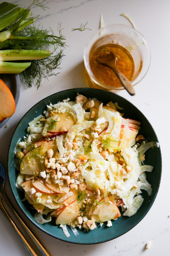 A fresh, vibrant apple salad sits in a green bowl, featuring thinly sliced fennel, apple slices, potato pieces, crumbled feta cheese, and hazelnuts. To the side is a glass jar containing a vinaigrette dressing and a spoon. Fresh fennel bulbs and sprigs are also partially visible.