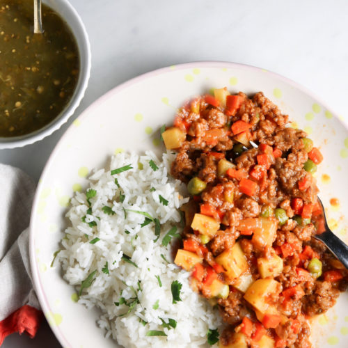 A plate filled with white rice garnished with chopped herbs and a serving of plant-based ground meat cooked with diced vegetables such as carrots and potatoes. A spoon rests in the meat portion. Next to the plate, there is a small bowl of green sauce with a spoon in it. A folded napkin is partially visible.