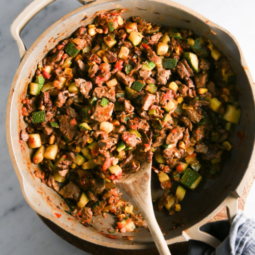 A large skillet filled with a colorful mixture of cooked vegetables and meat resembles Calabacitas con Carne. Ingredients include cubed zucchini, corn, tomatoes, and pieces of beef. A wooden spoon rests in the skillet, and a grey cloth is tucked underneath on a marble countertop.
