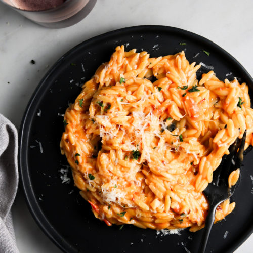 A black plate with creamy orzo pasta topped with grated cheese and herbs. The tomato-based sauce has a touch of spicy flavor, garnished with chopped parsley. A black fork lies on the plate, and a glass of red wine is partially visible at the top left corner of the image. A light gray cloth is placed to the left.