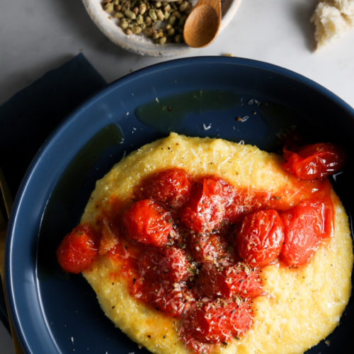 A blue bowl holds a serving of creamy polenta topped with blistered cherry tomatoes and a sprinkle of grated cheese and herbs. Next to the bowl are a small dish of salt with a wooden spoon and a few pieces of crusty bread on a marble surface.