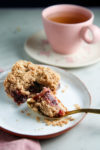 A piece of crumbly plum streusel dessert with a golden spoon rests on a white plate. Part of the dessert is scooped out, revealing a dark berry filling. In the background, a pink cup with tea sits on a matching saucer on a light-colored surface. Some crumbs are scattered around the plate.